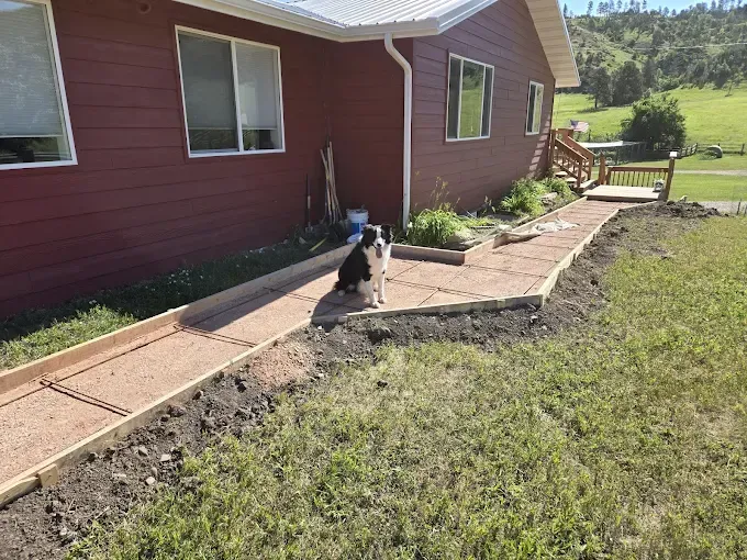 Dog sits on a concrete walkway next to a red house under construction, grass lawn surrounds.