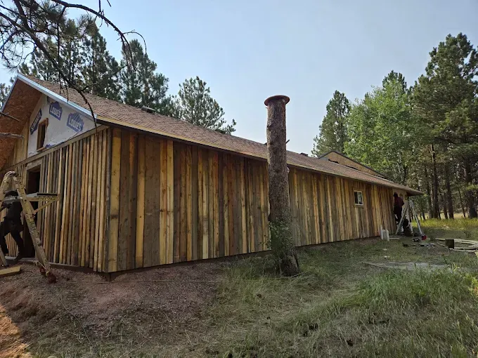 Cabin with wood siding under construction in a forested area; cedar shake roof, chimney.