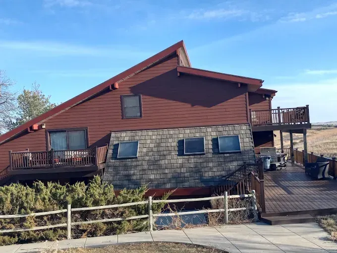 Red, two-story house with deck, shingled base, and sloped roof on a sunny day.