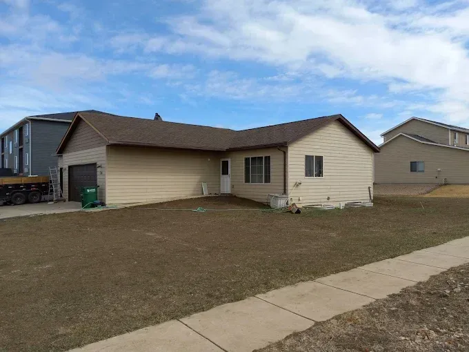 Tan house with brown roof, garage, and lawn under a blue sky with clouds.