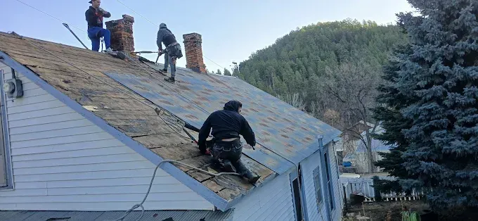 Roofers work on removing old shingles from a house roof with brick chimneys and a mountain background.