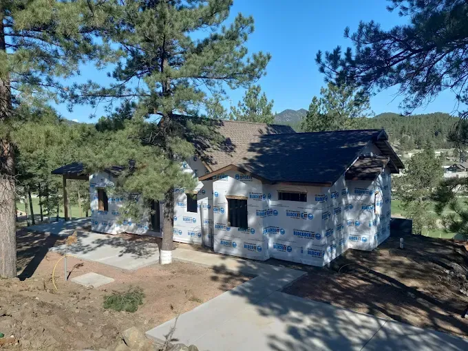 House under construction, wrapped in blue protective covering, surrounded by trees and a paved driveway.