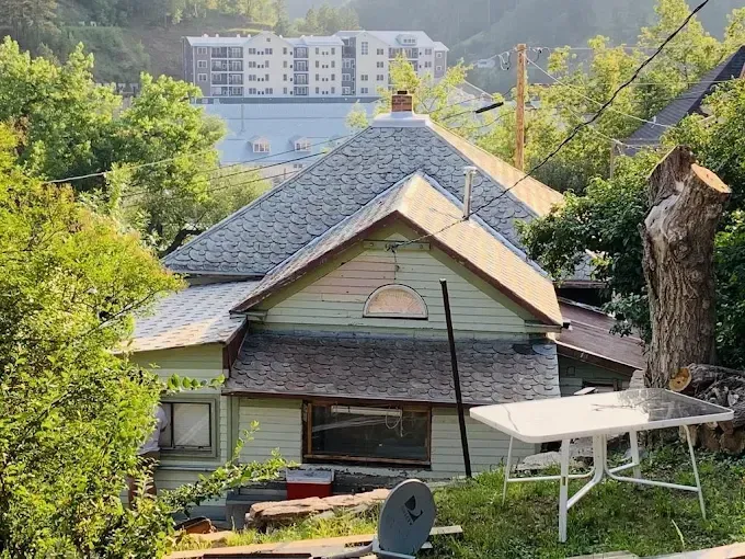 Green house with gray roof, in a hillside setting with trees. A white table is in the yard.