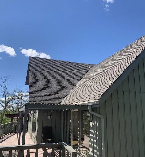 A house with gray shingles and green siding under a blue sky. A deck is in the foreground.
