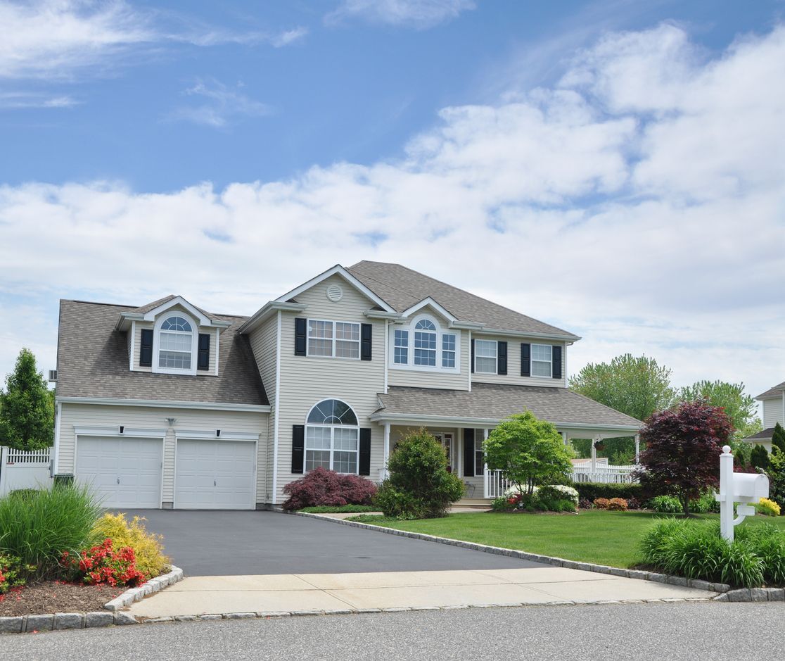 Two-story house with light siding, gray roof, two-car garage, and landscaped yard under a blue sky.