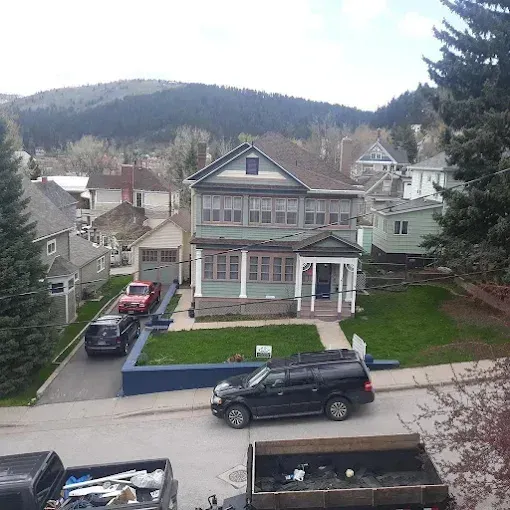 Two-story green house with porch and cars parked on the street, set in a mountain town.