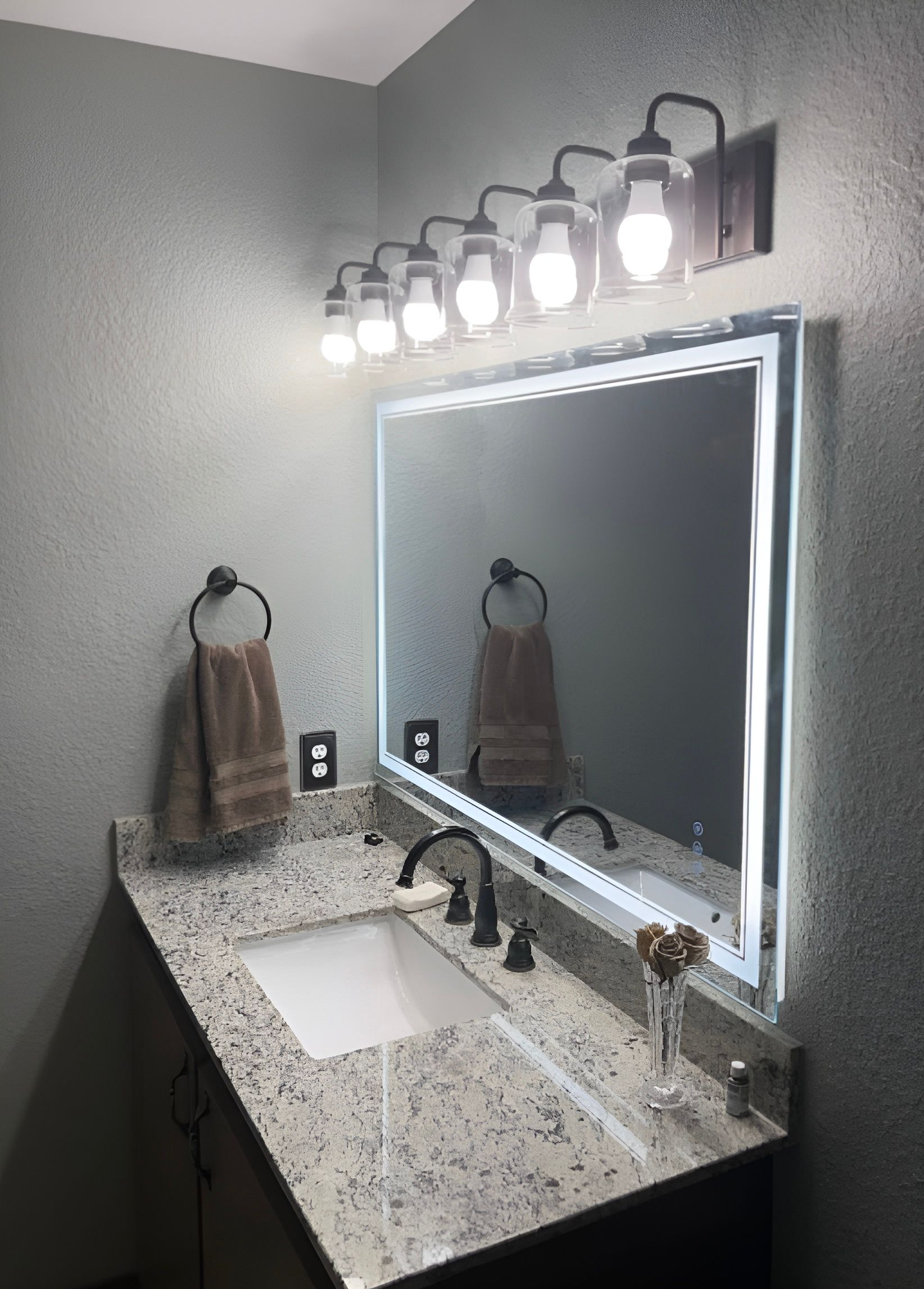 Modern bathroom vanity with lit mirror, speckled countertop, sink, towel, and wall-mounted light fixture.