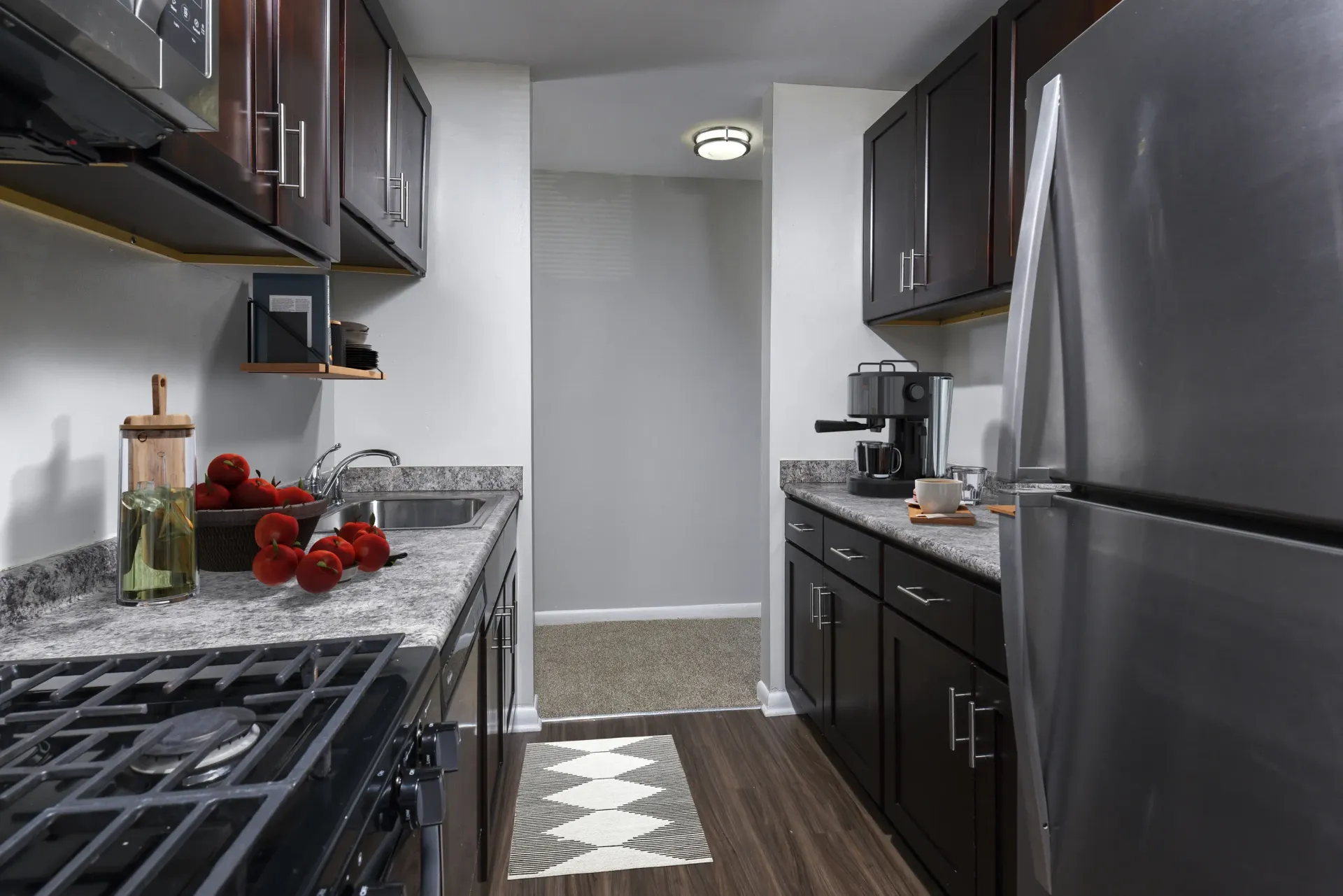 Small galley kitchen with dark wood cabinets, stainless steel fridge, and granite-like countertops at Holly Court, offers apartments for rent in Pitman, NJ.