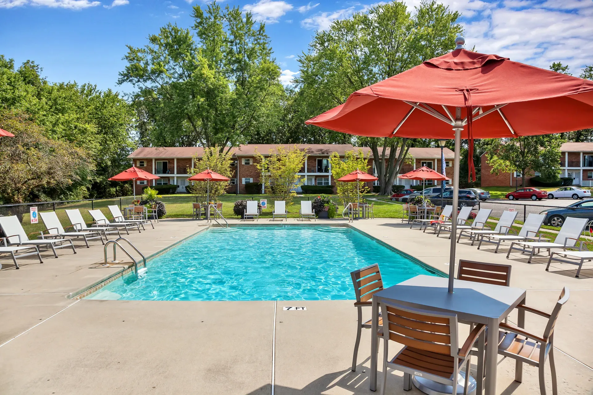 Outdoor apartment pool area with lounge chairs, tables under red umbrellas, and trees at Holly Court, offers apartments near downtown Pitman, NJ.