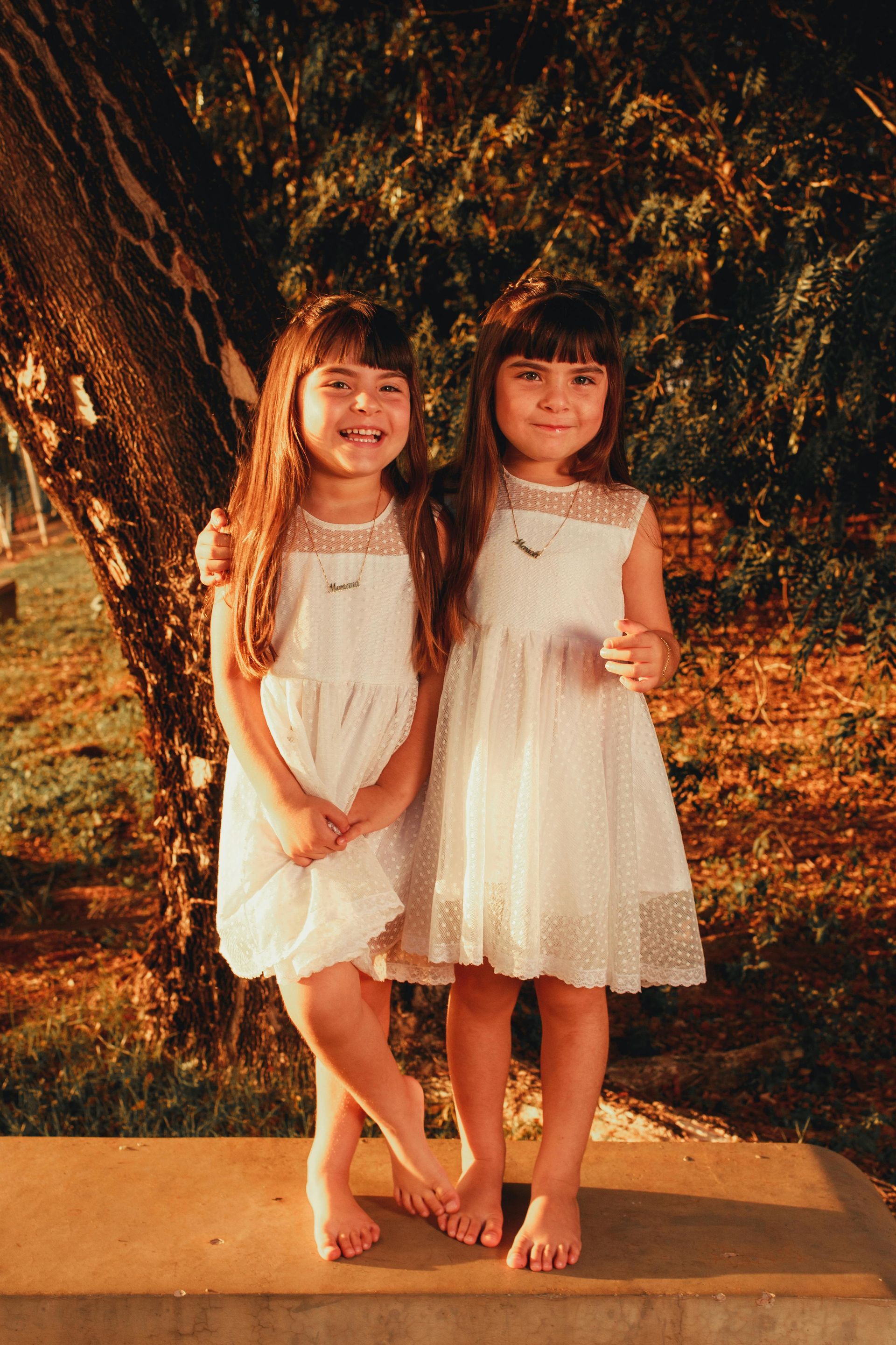 Two young girls in white dresses smile, barefoot, standing outdoors with a tree in the background.