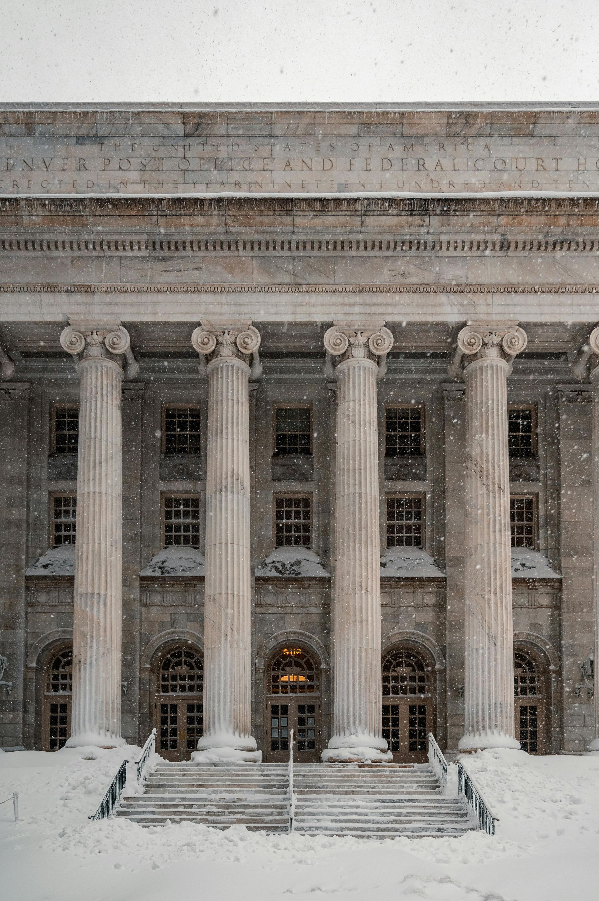 Snowy neoclassical building facade with tall columns and a lit entrance at the top of steps