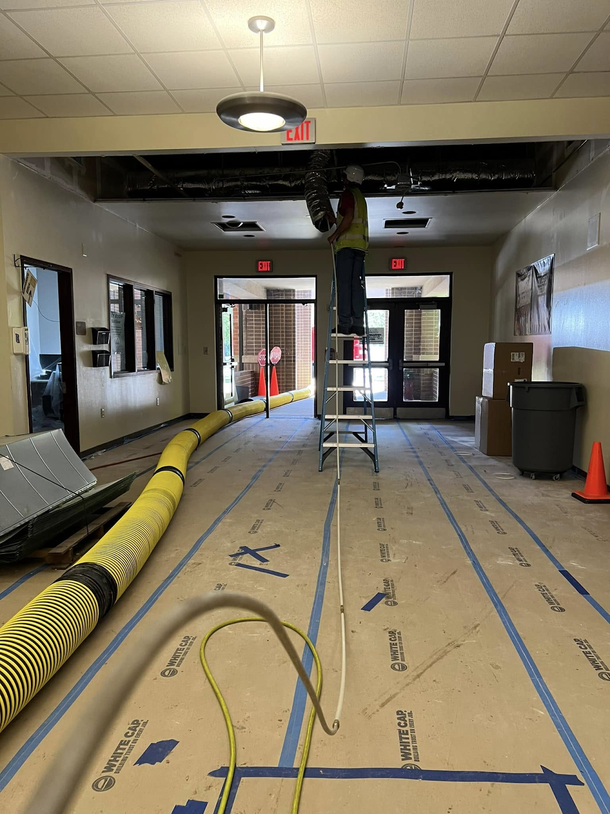 Person on ladder working on ceiling; hallway under construction; yellow hose on floor.