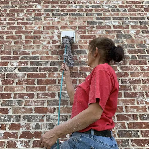 Woman cleaning a dryer vent on a brick wall with a long-handled brush.
