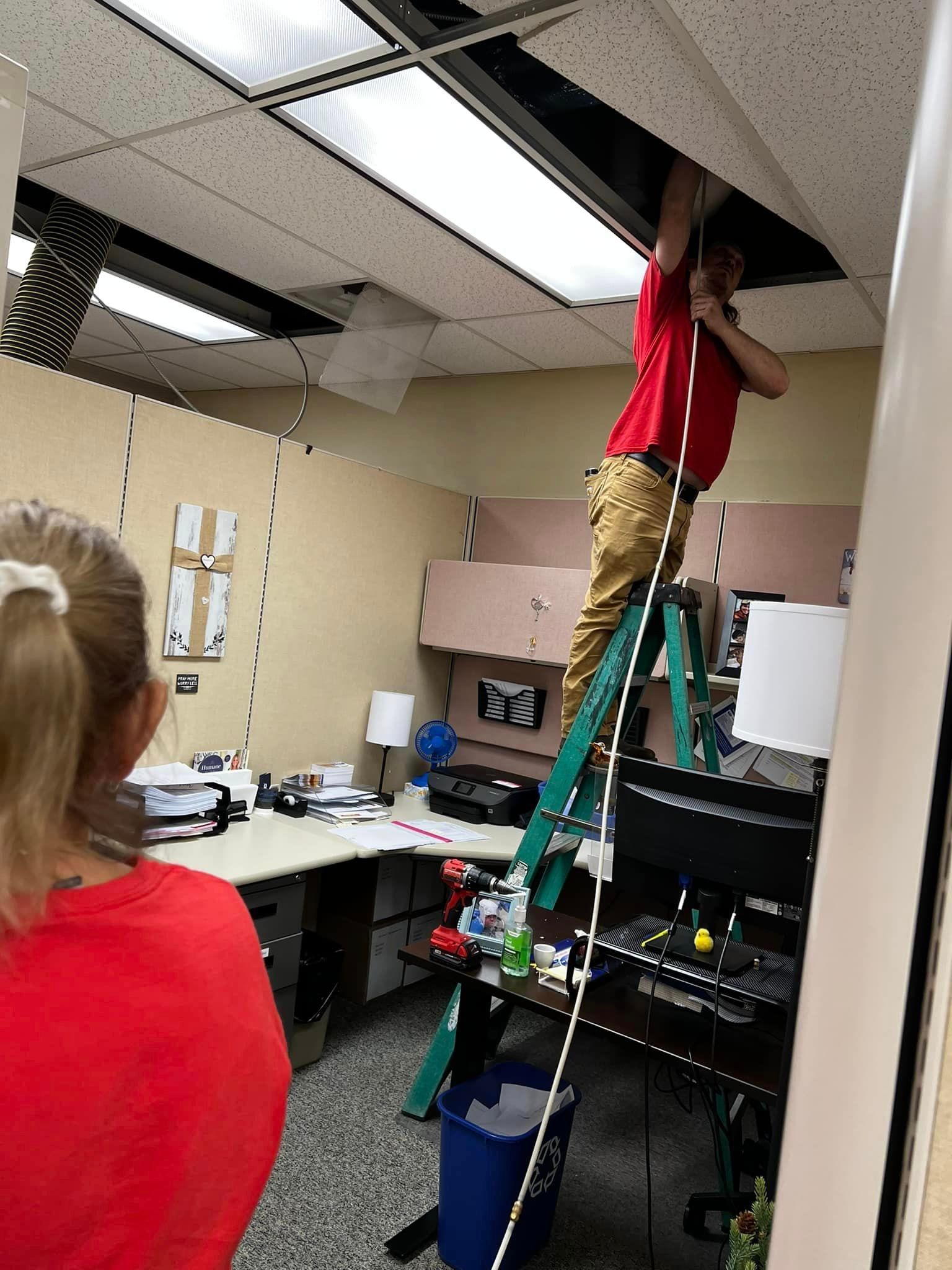 A man in a red shirt is standing on a ladder in an office .