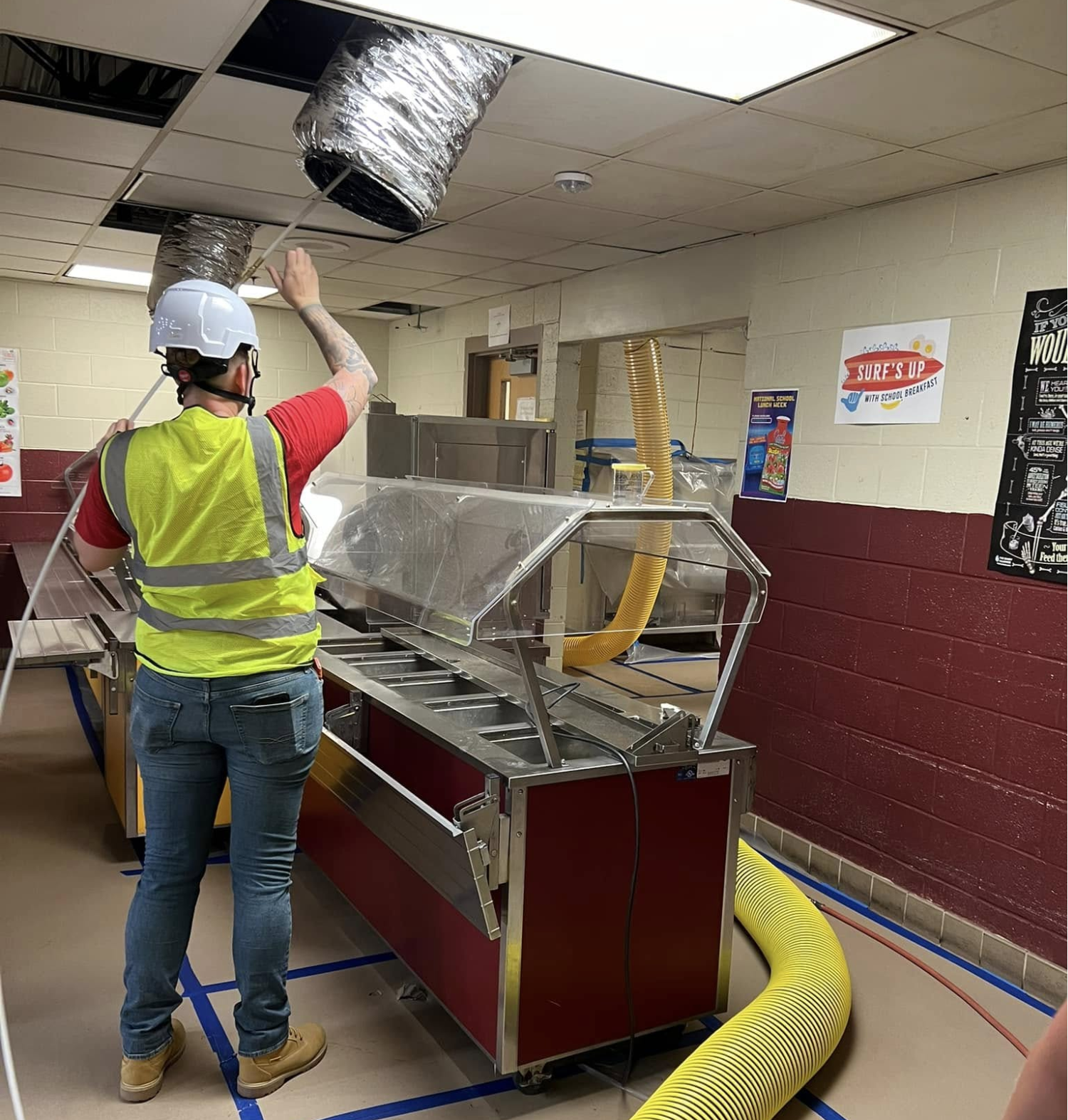 A worker in a hard hat and high-visibility vest pulls a flexible vent hose from a ceiling grid above a food service line.