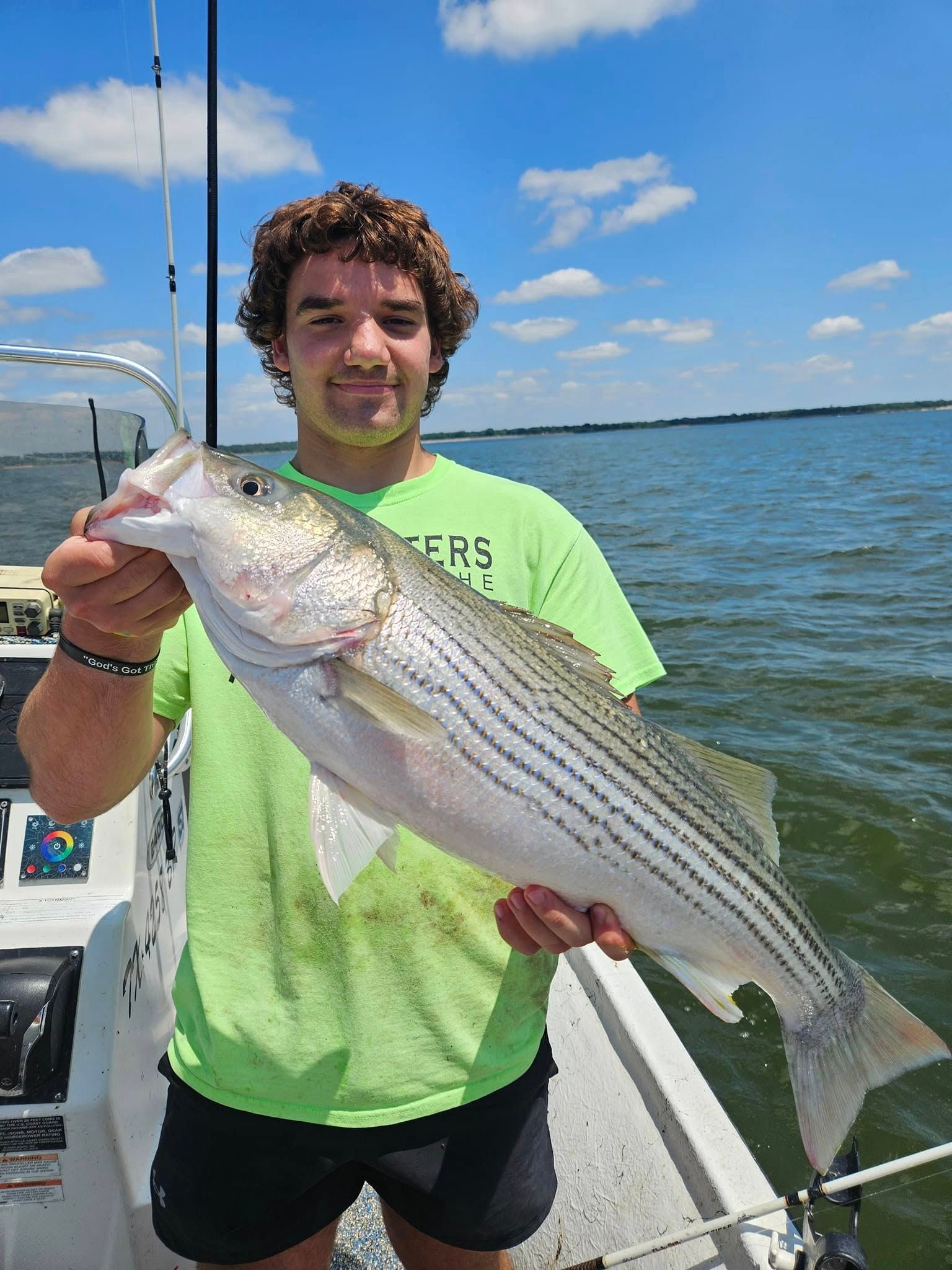 Man on a boat smiles, holding a striped bass. Blue sky, water. Green shirt.