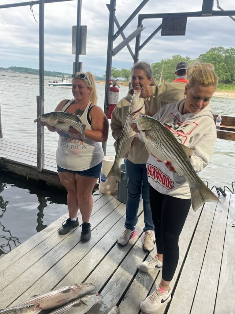 Three women on a dock holding up striped bass they caught. Water, cloudy sky in the background.