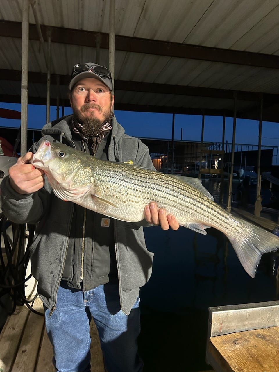 Man holding a large striped bass. He wears a hat, jacket, and jeans, by a dock at dusk.