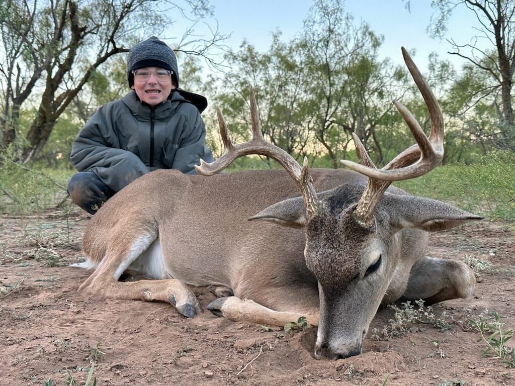 Boy kneeling next to a large buck with antlers on the ground. Boy smiles, wearing a jacket and hat.