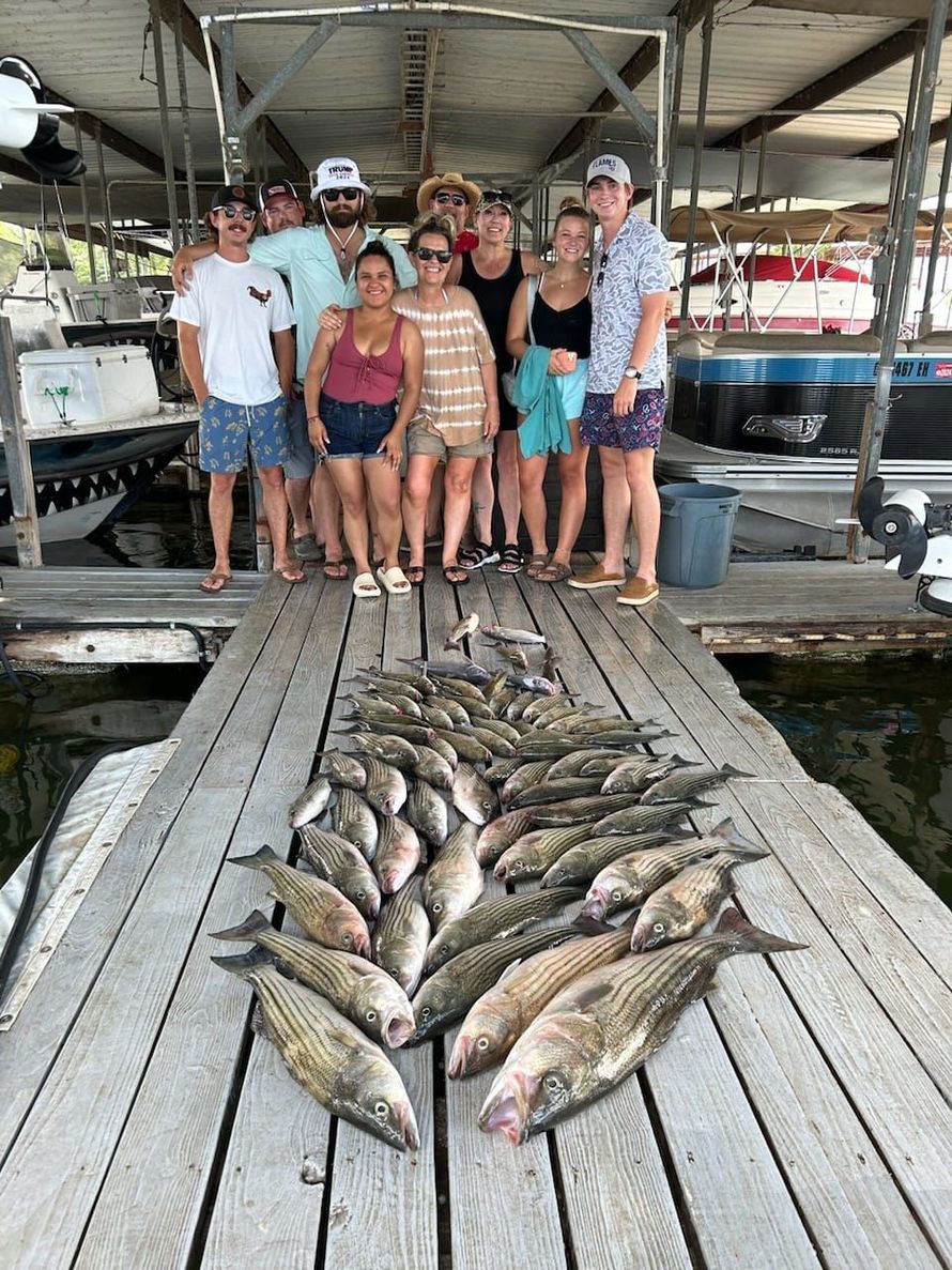 Group of people on a dock with a large pile of striped fish, under a boathouse.