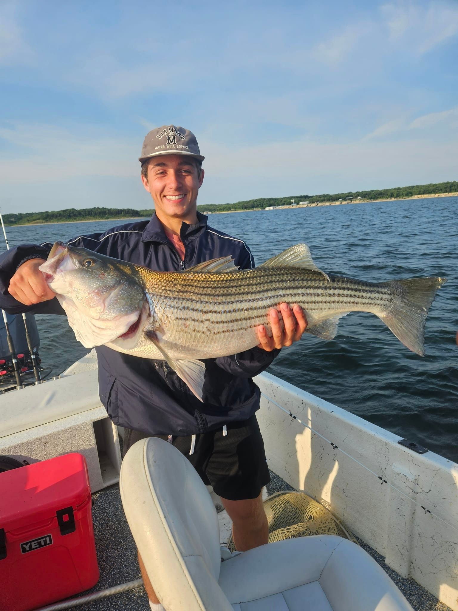 Man on boat holds up a striped bass. He's smiling in front of water and a treeline.