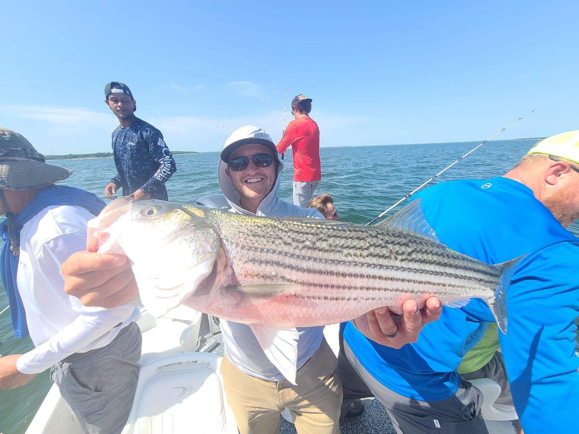 Man on a boat holds a large striped bass he caught. Blue water and sky. People fishing.