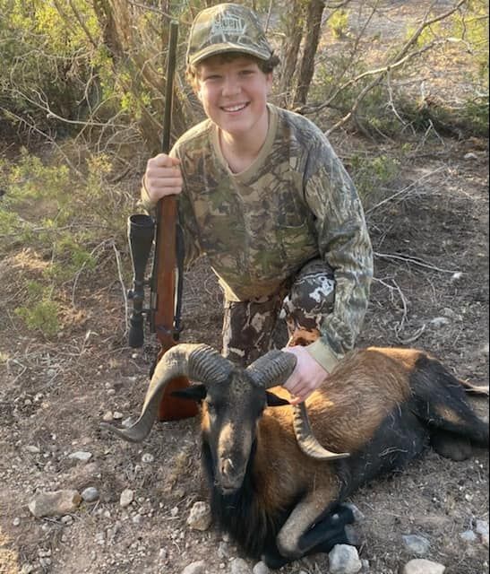 Young hunter kneels with a rifle next to a dead ram in a desert setting; he smiles.