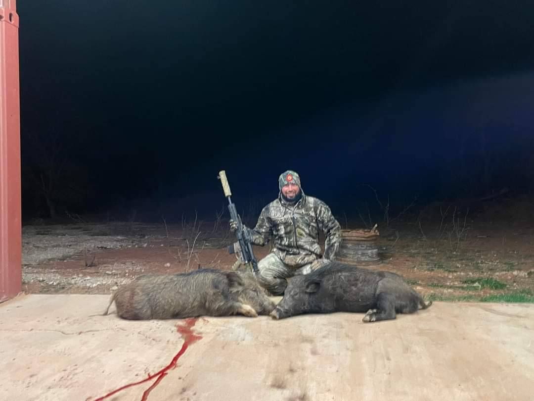 Hunter kneels beside two dead wild boars on a concrete pad at night; holds a rifle.