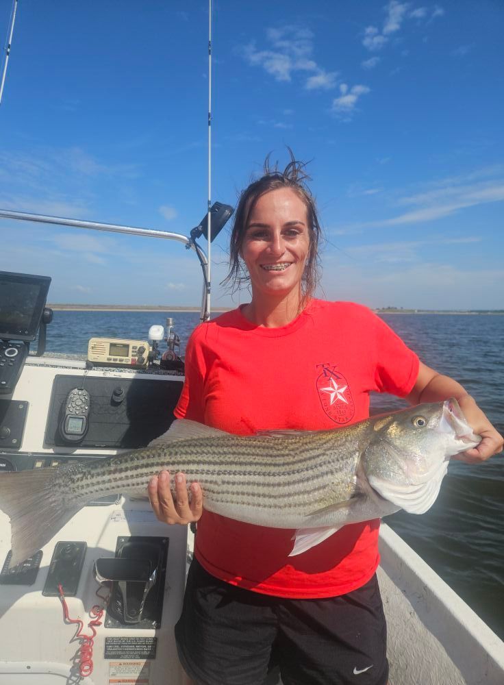 Woman on a boat holding a striped bass fish, smiling. Sunny day, blue sky and water.