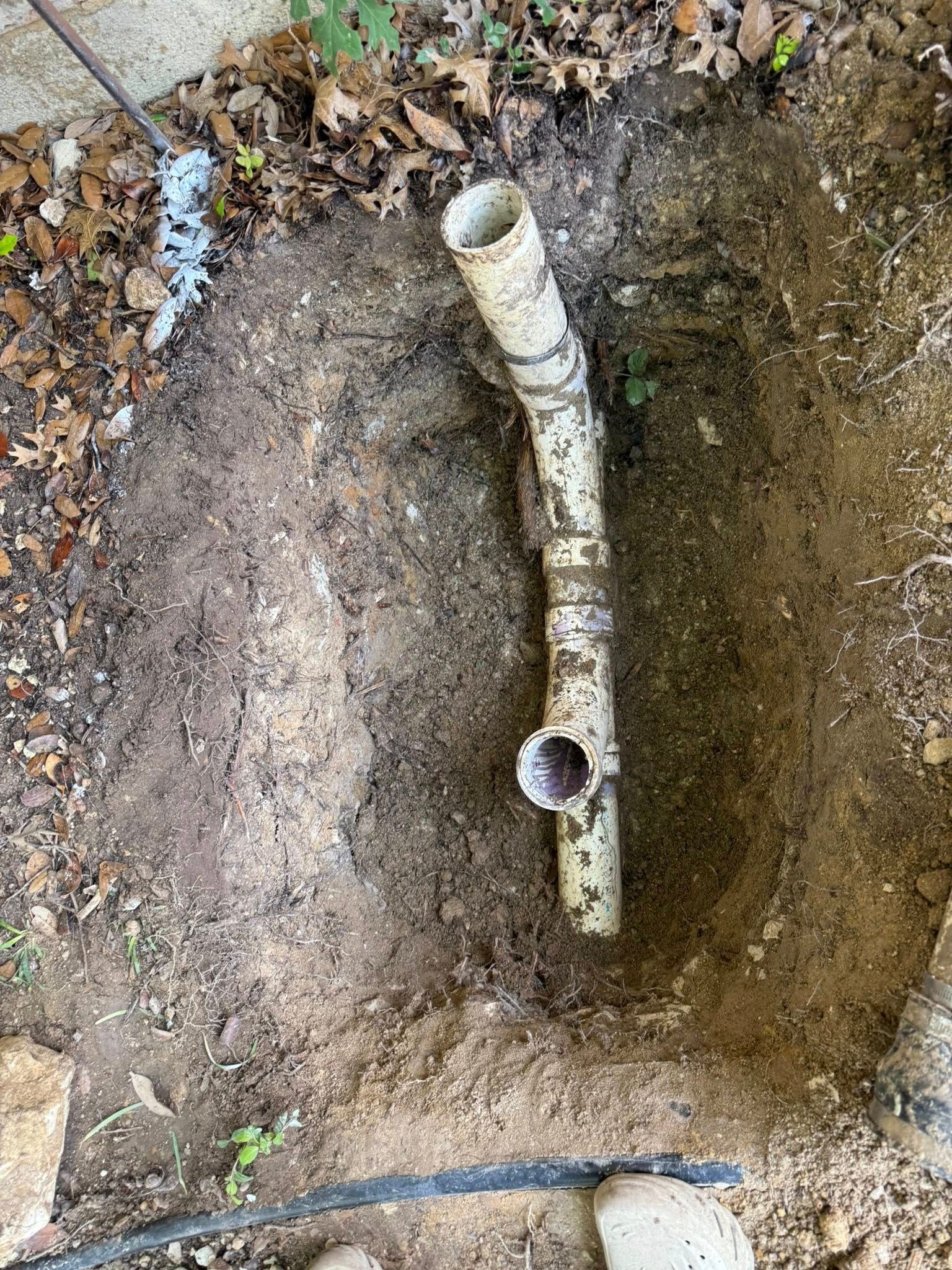 A white PVC pipe assembly with a T-junction connector, partially exposed in an excavated dirt ground.
