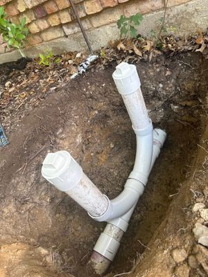 A dual-riser white PVC plumbing cleanout assembly installed in a dirt trench next to a brick wall.