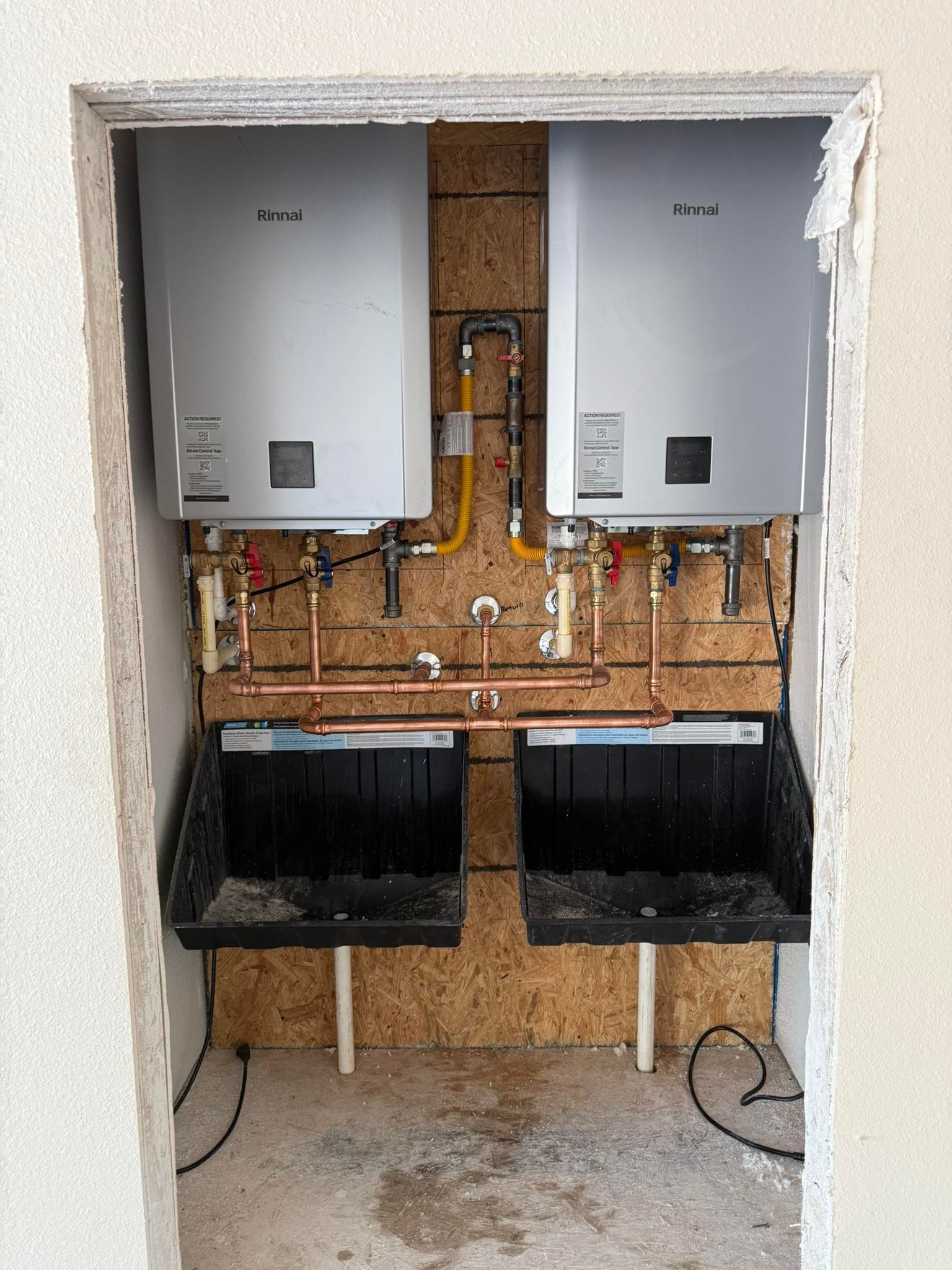 Two wall-mounted tankless water heaters installed in an unfinished alcove with copper plumbing and black drip trays.