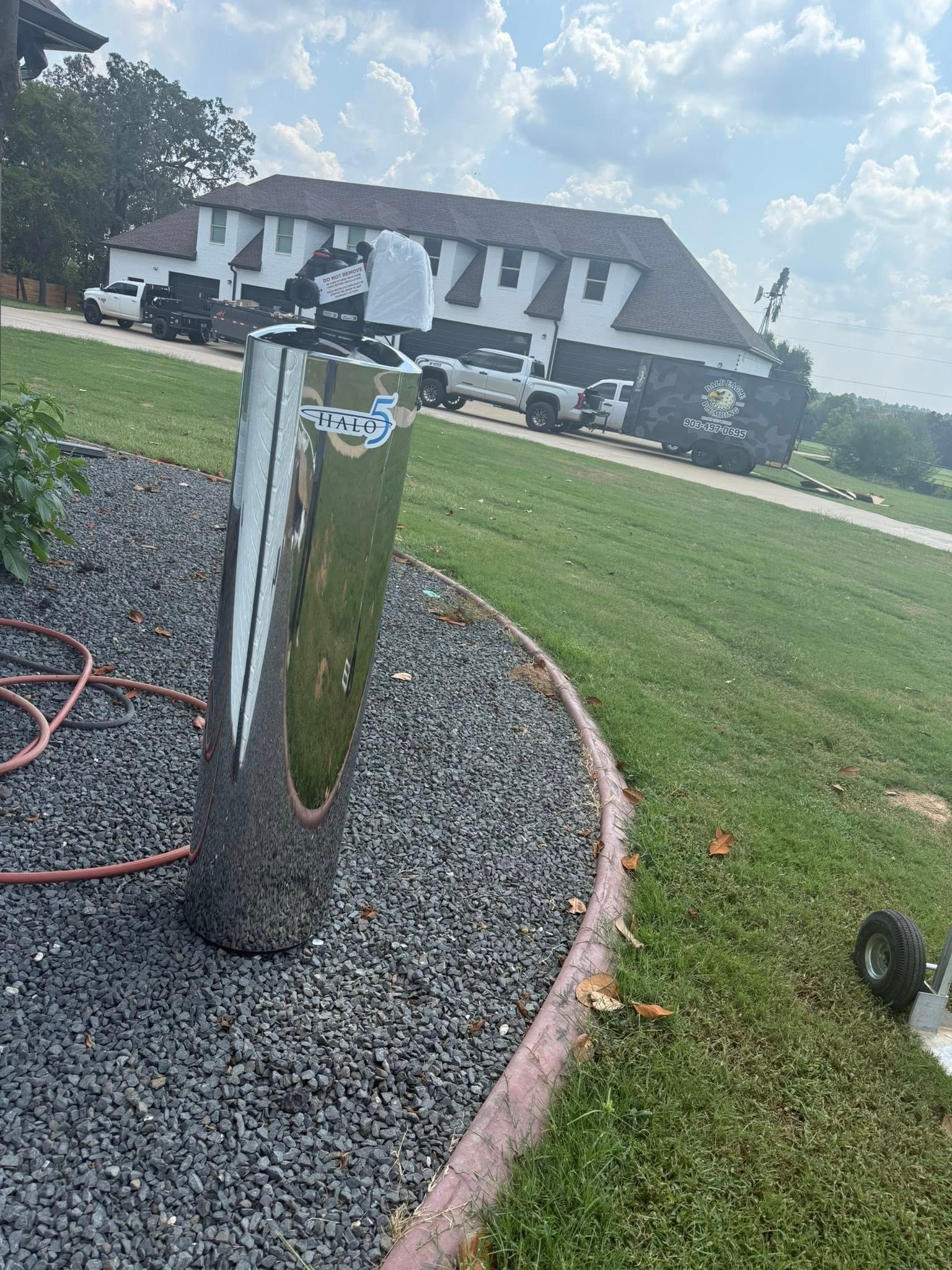 A reflective metal post stands in a dark gravel bed, with a house, trucks, and a large black dumpster in the background.