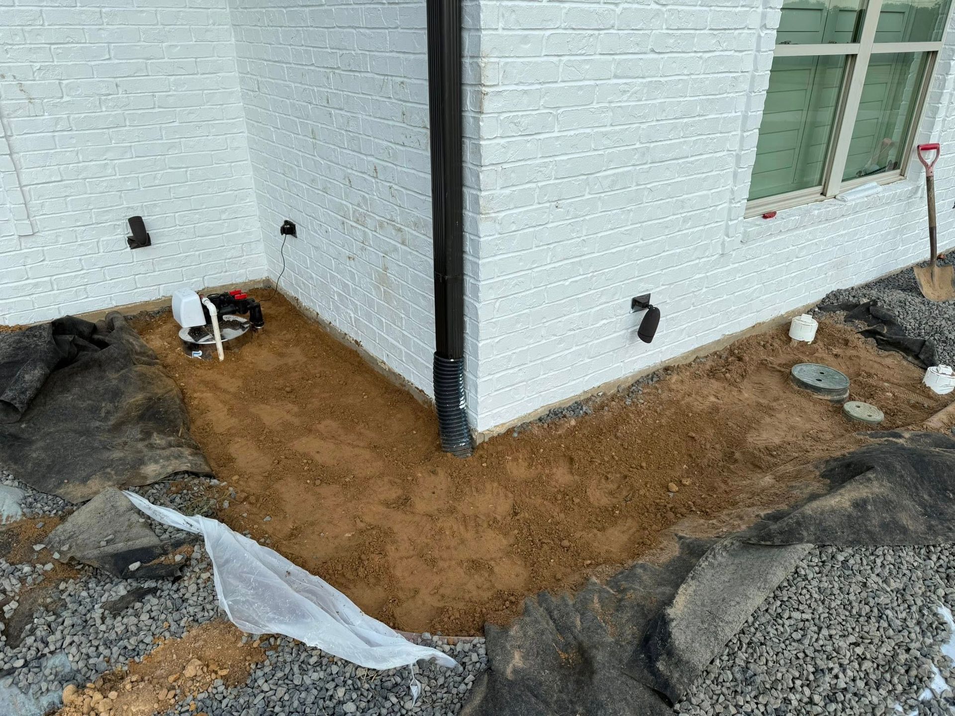 An exterior corner of a white brick house with a dark downspout, sand-filled foundation bed, and landscaping materials.