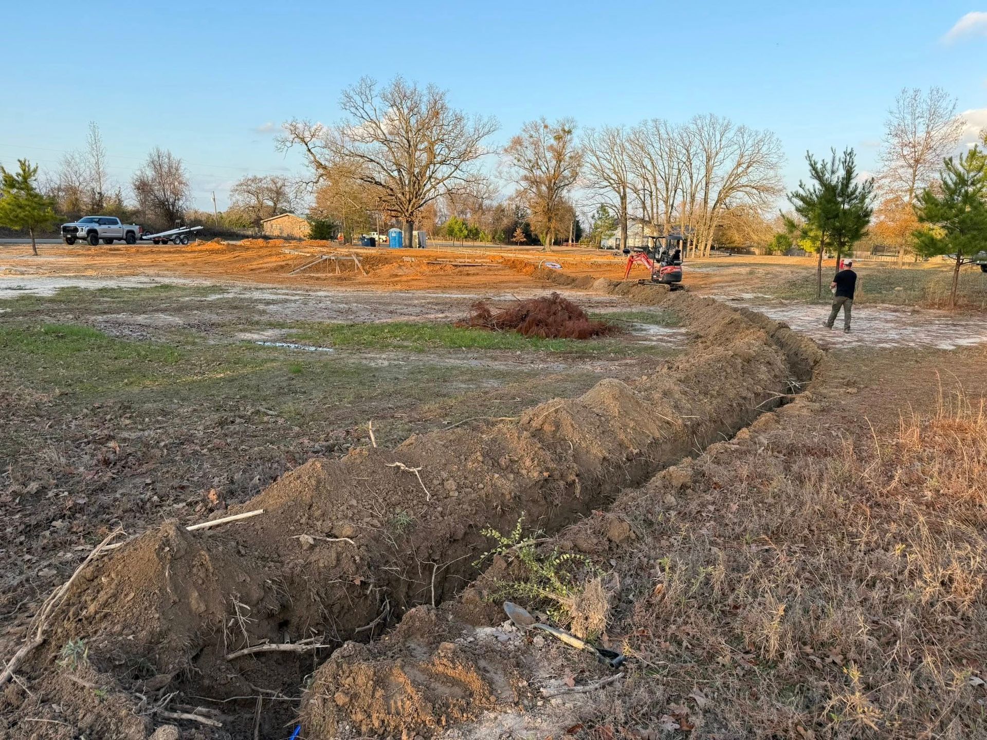 A trench dug through a grassy, sunlit field with a person standing nearby and construction machinery in the distance.