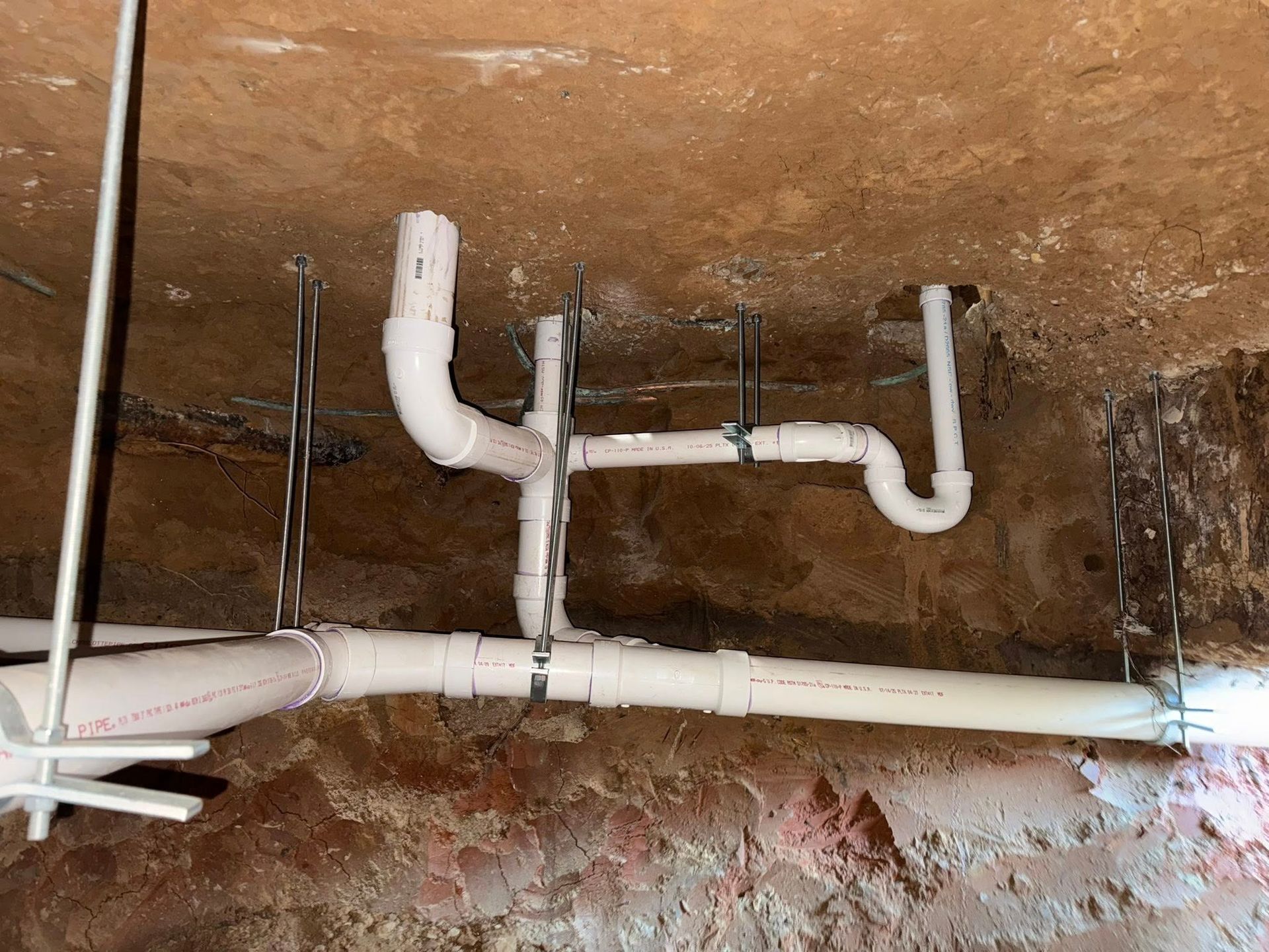 White PVC plumbing pipes are suspended from a dirt ceiling using metal straps and threaded rods in a crawlspace.