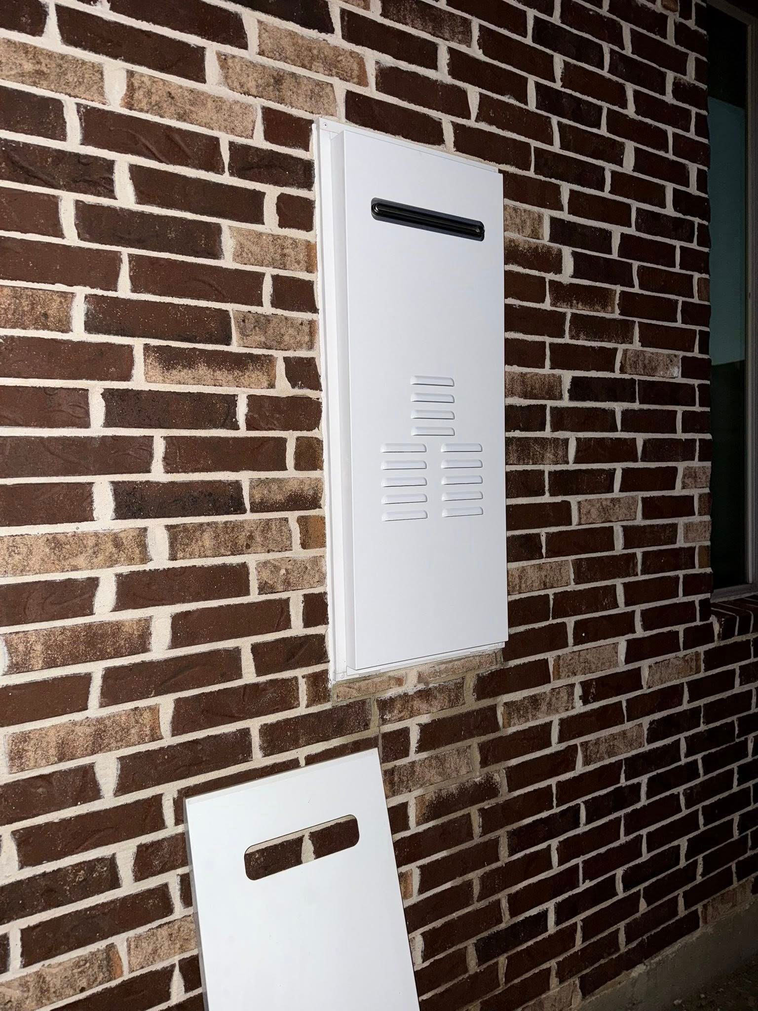 A white rectangular tankless water heater cover mounted on a brown brick wall with a detached panel lying below it.