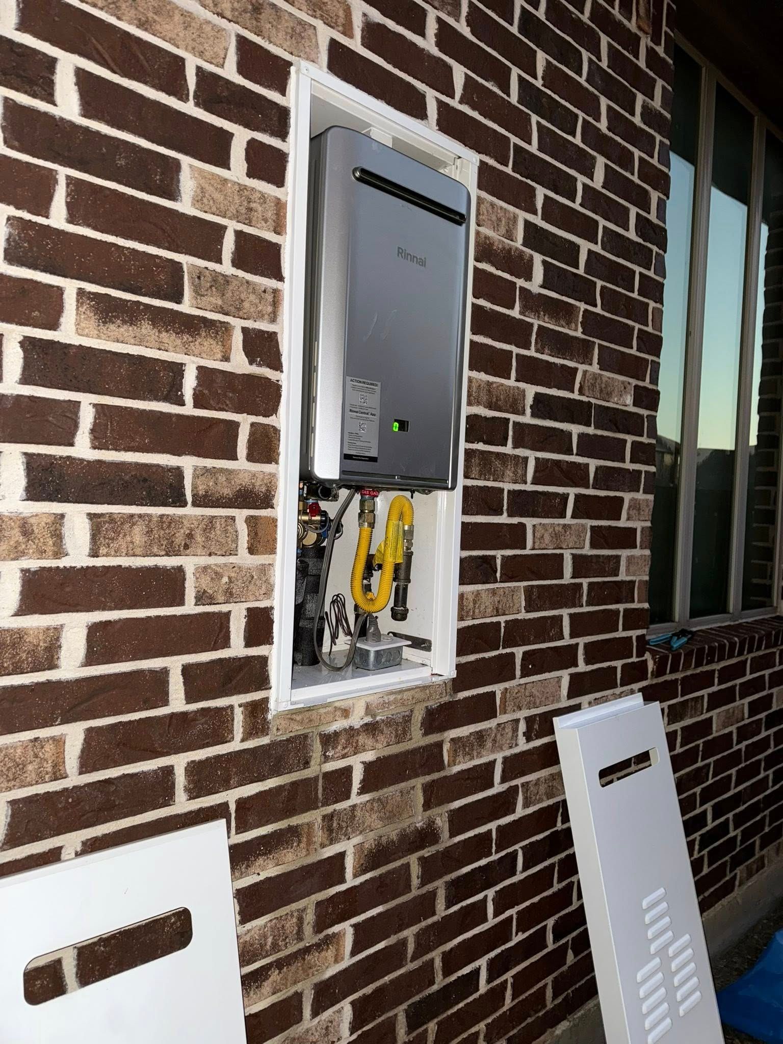 A tankless water heater installed inside a white recessed wall box on an exterior brick wall.