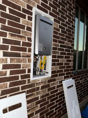A tankless water heater installed in an exterior brick wall recess, with its cover panel resting on the ground.
