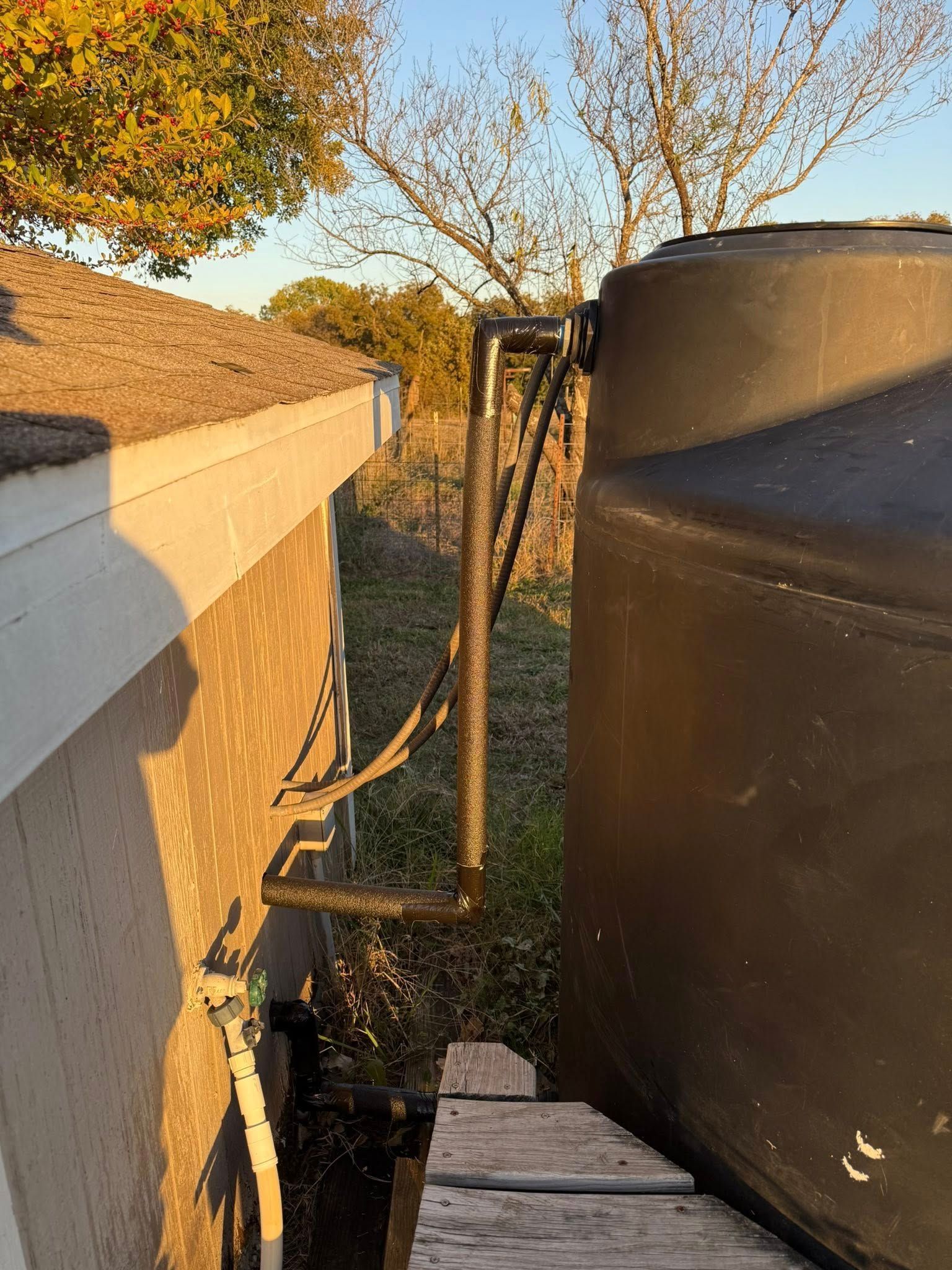 A black plastic water storage tank stands next to a building wall, connected by a vertical black pipe and plumbing.