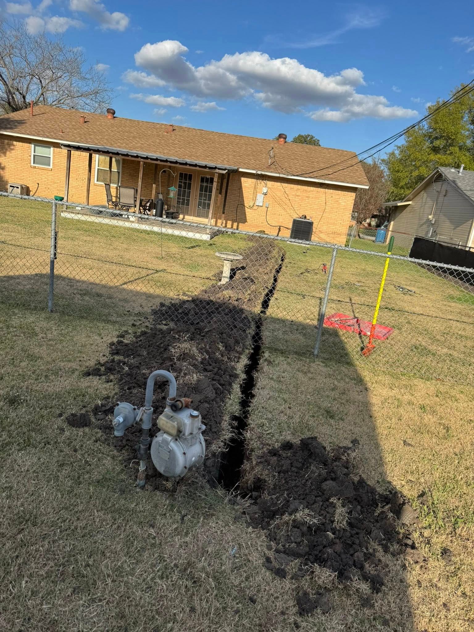 A trench cut through a residential lawn leading toward a brick house, with a gas meter visible in the foreground.