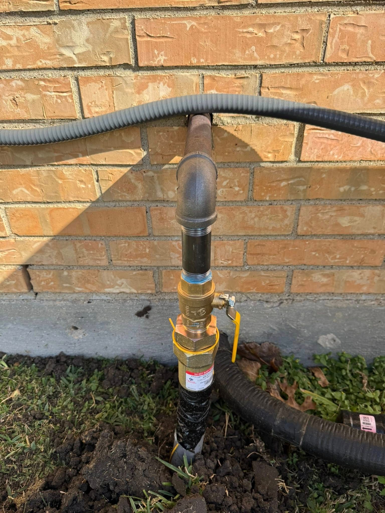 A yellow-handled gas shut-off valve installed on a pipe against a brick wall, with flexible conduit attached.