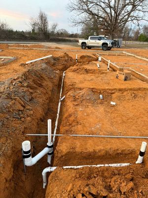 White plumbing pipes installed in trenches at a construction site with orange soil and a pickup truck in the distance.