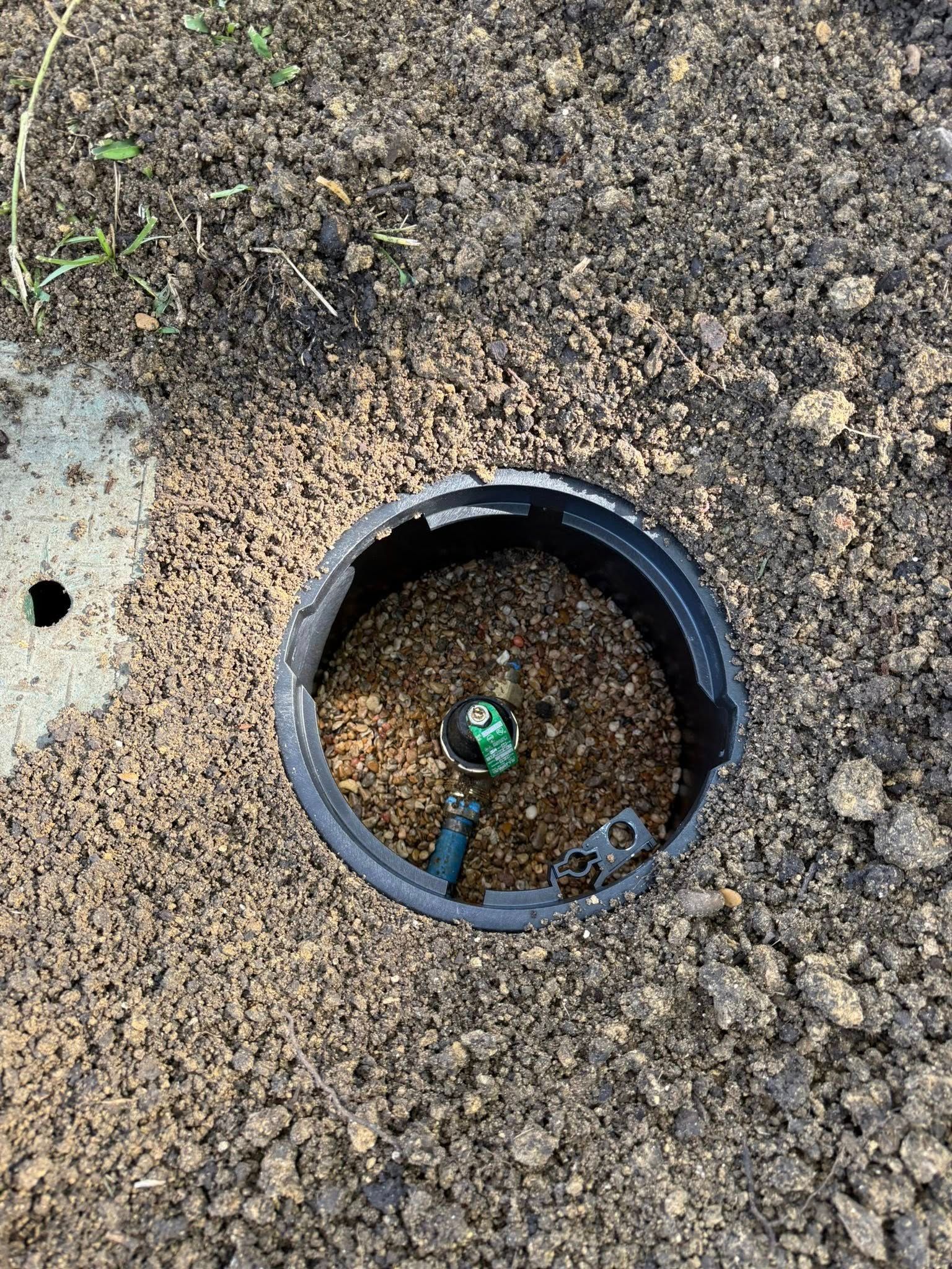 A round irrigation valve box embedded in the ground, showing a green sprinkler head and connection pipes inside.