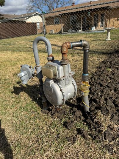 A residential natural gas meter with a regulator and piping, installed in a grassy yard near a chain-link fence.