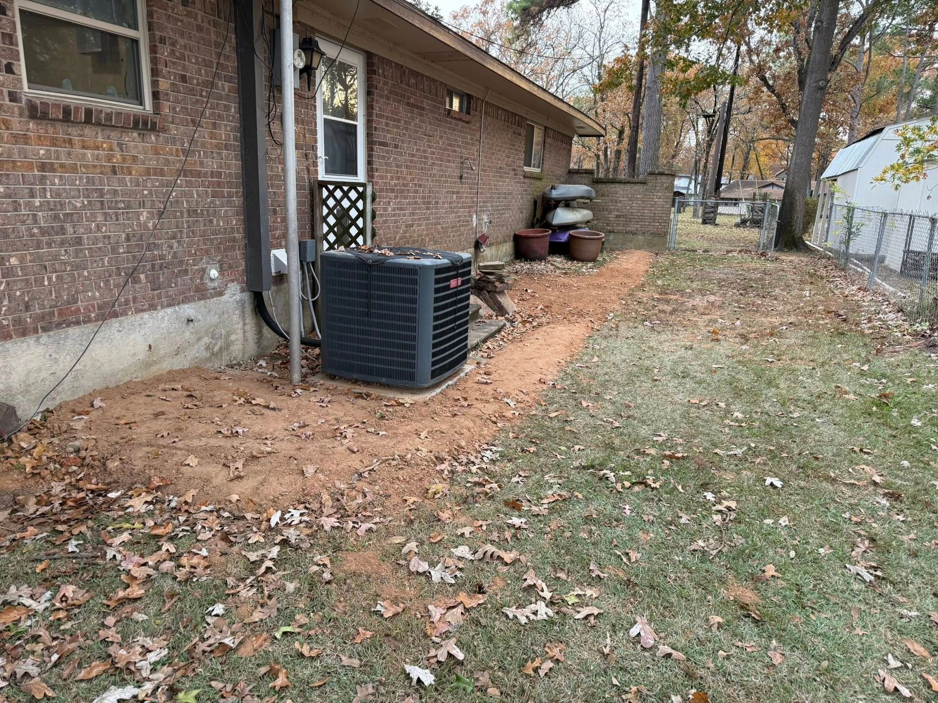 An air conditioning unit sits on a patch of loose dirt beside a brick house with a white window and surrounding yard.