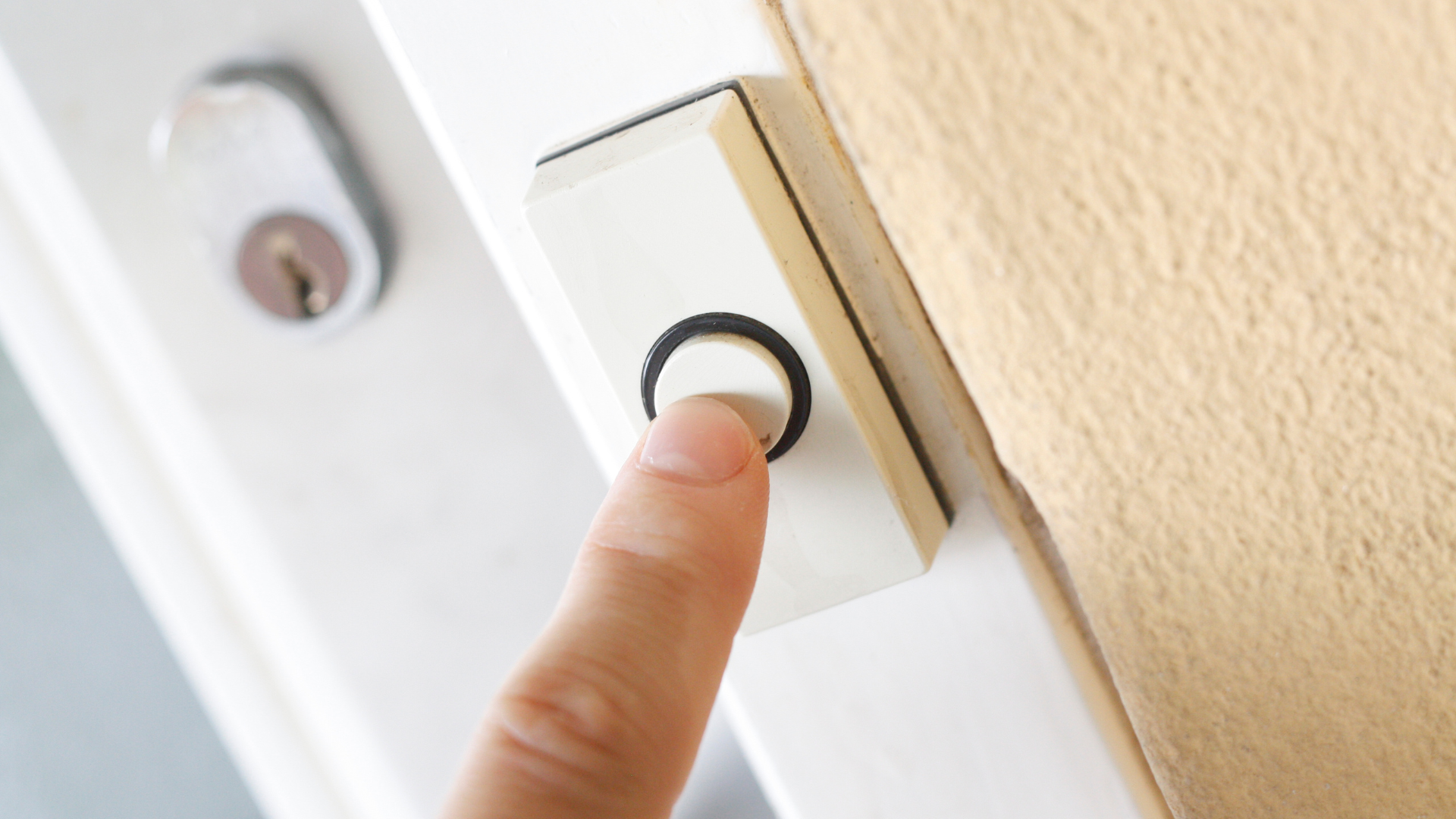 Finger pressing a white doorbell button on a white door frame next to a beige fabric.