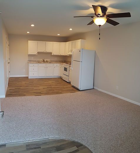 An empty living room with a kitchen and a ceiling fan.