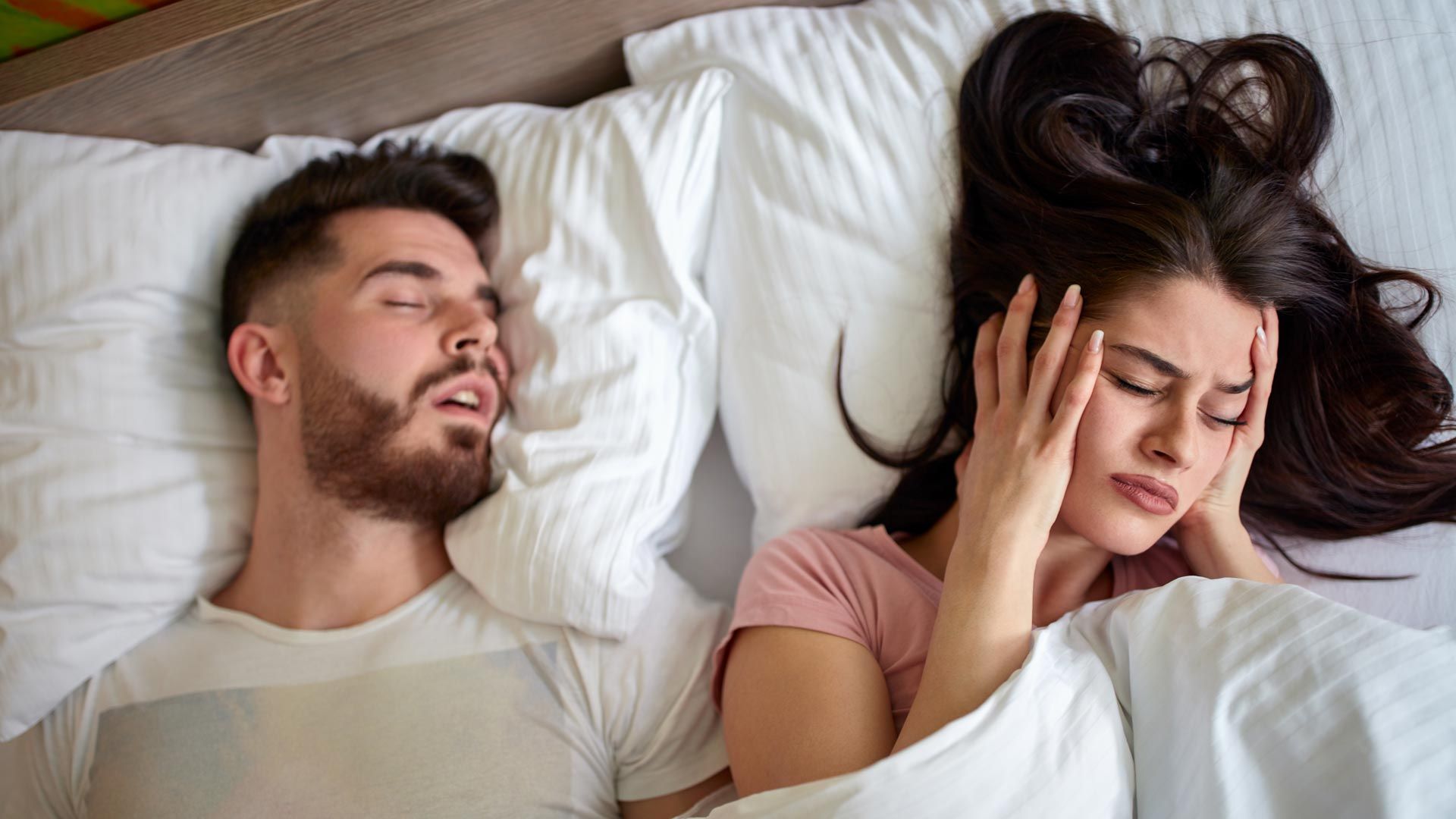 Frau hält sich im Bett Ohren zu, weil Mann neben ihr schnarcht.