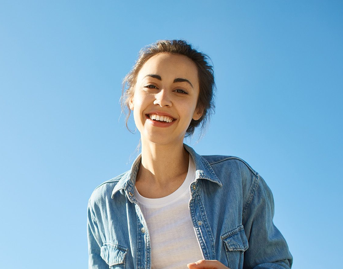 Woman smiling, blue sky background, wearing denim shirt and white tee.